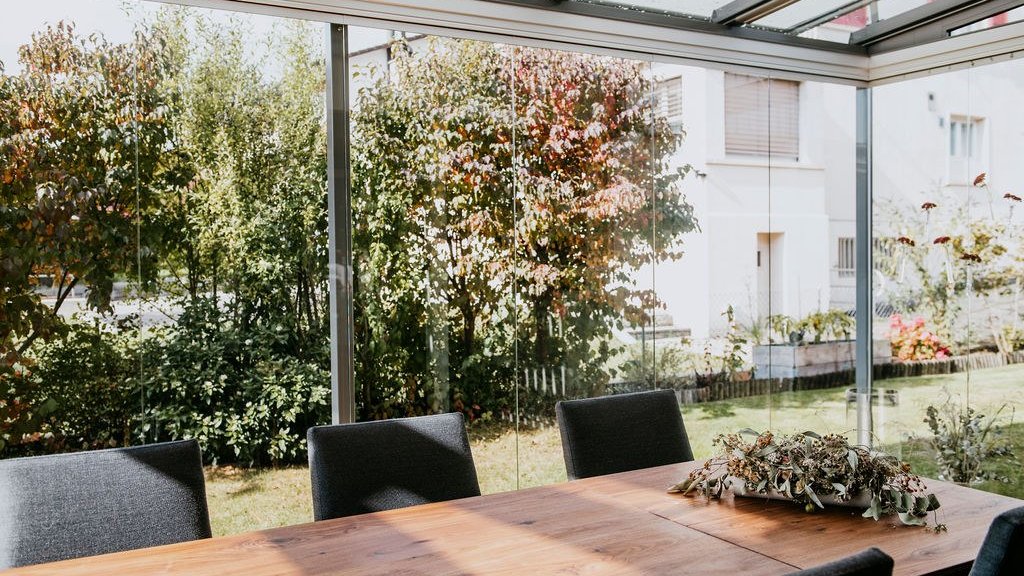 Esstisch im Sommergarten mit ROOFS Glasdach und COVER Schiebe-Drehverglasung vor herbstlichen Bäumen
