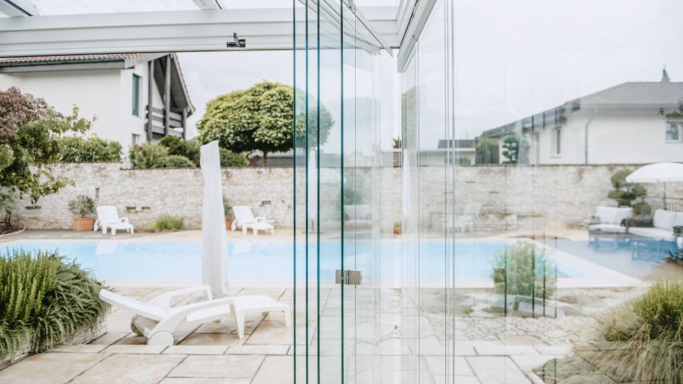 Jardin d’été avec vitrage COVER partiellement ouvert, chaise longue et piscine en arrière-plan